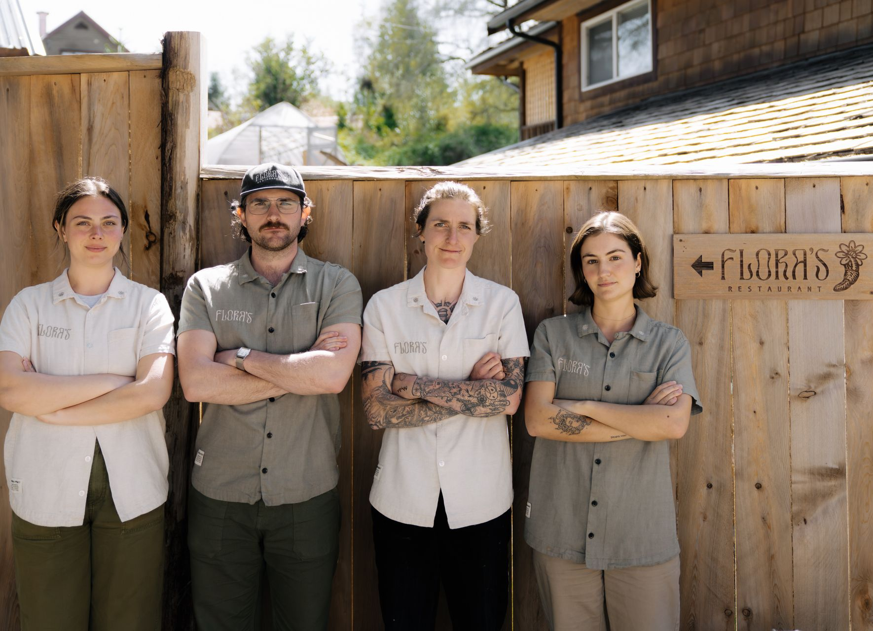 Four people in casual, light-colored shirts stand with arms crossed against a wooden fence with a small sign for Flora’s.