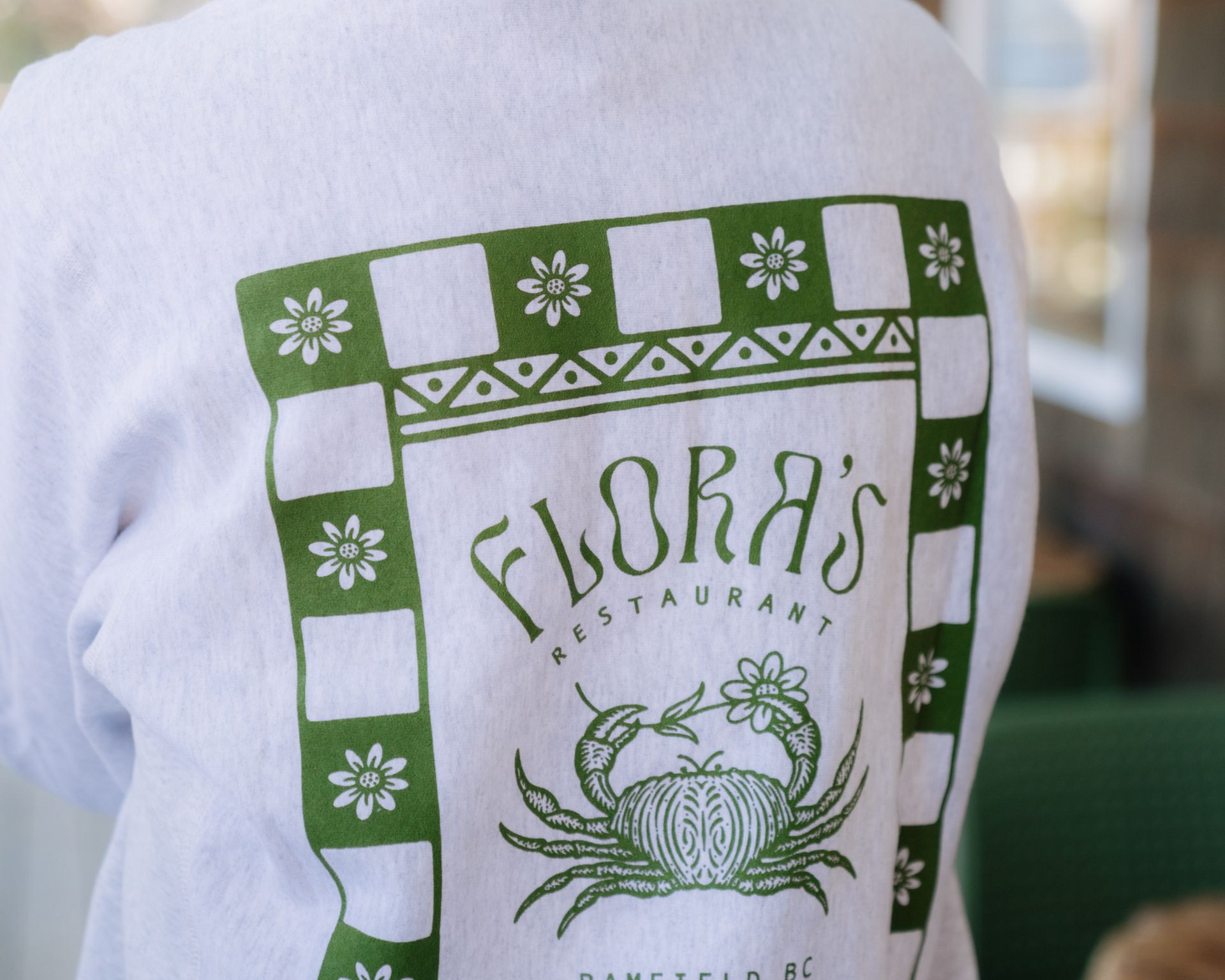 A green screen-printed graphic on a white sweatshirt showing a crab, flowers, and the text 