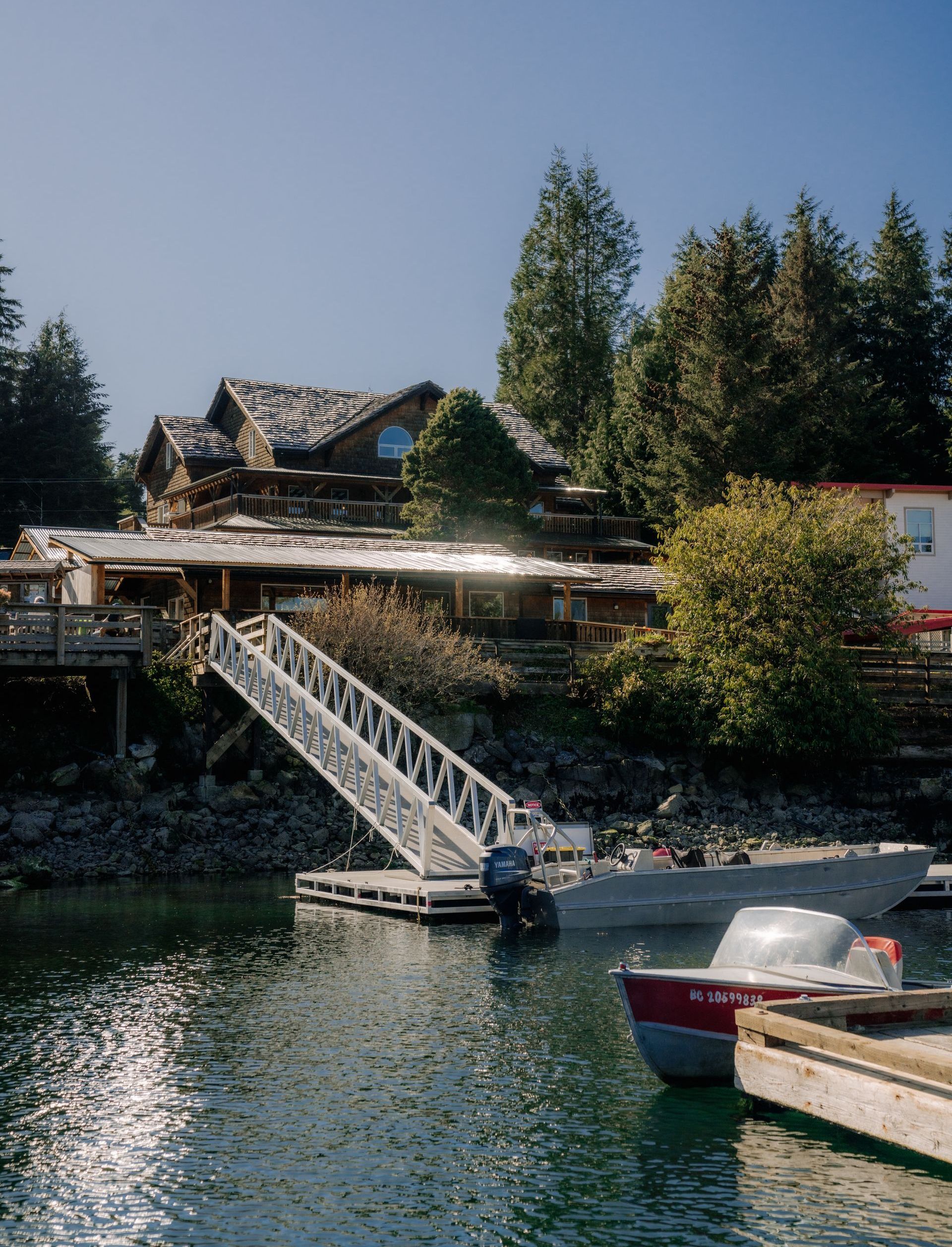 A rustic wooden lodge on a wooded shoreline, connected to a floating dock by a long metal ramp, with boats anchored nearby.