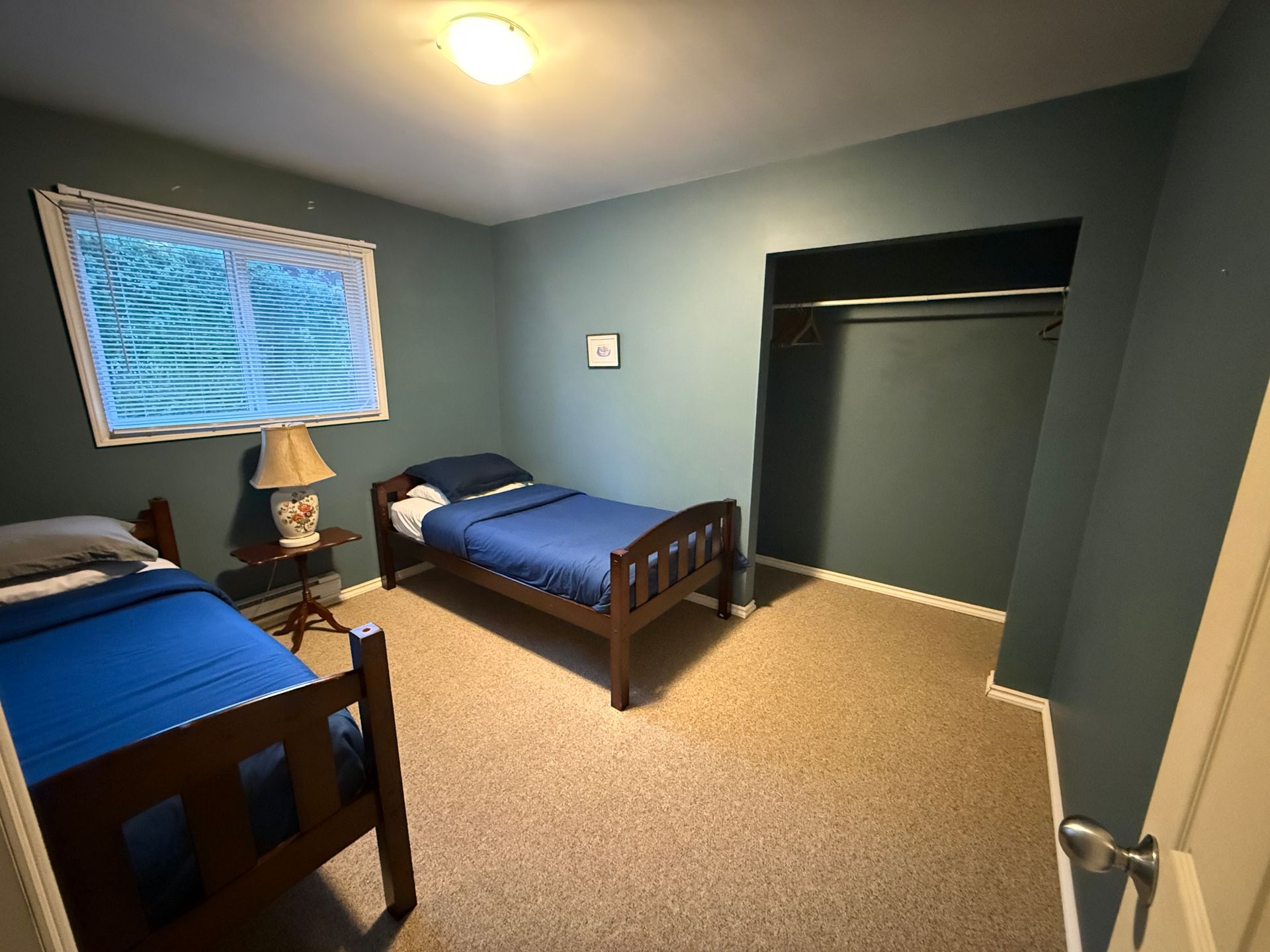 Bedroom with blue walls, two twin beds with blue bedding, a lamp on a small table, and an open closet.