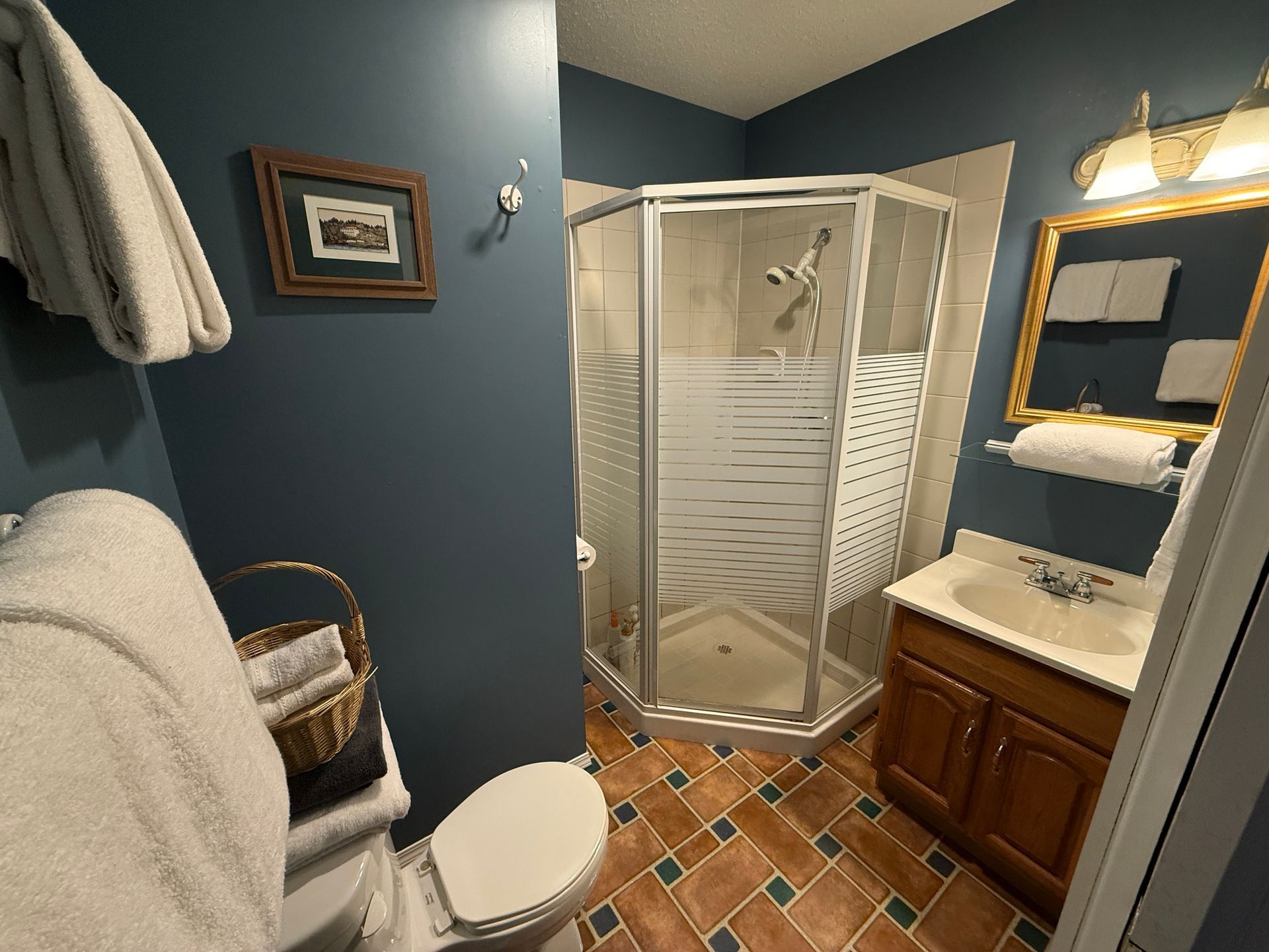 A bathroom with dark blue walls, a corner shower, wood vanity, and brown tiled floor with decorative green inserts.