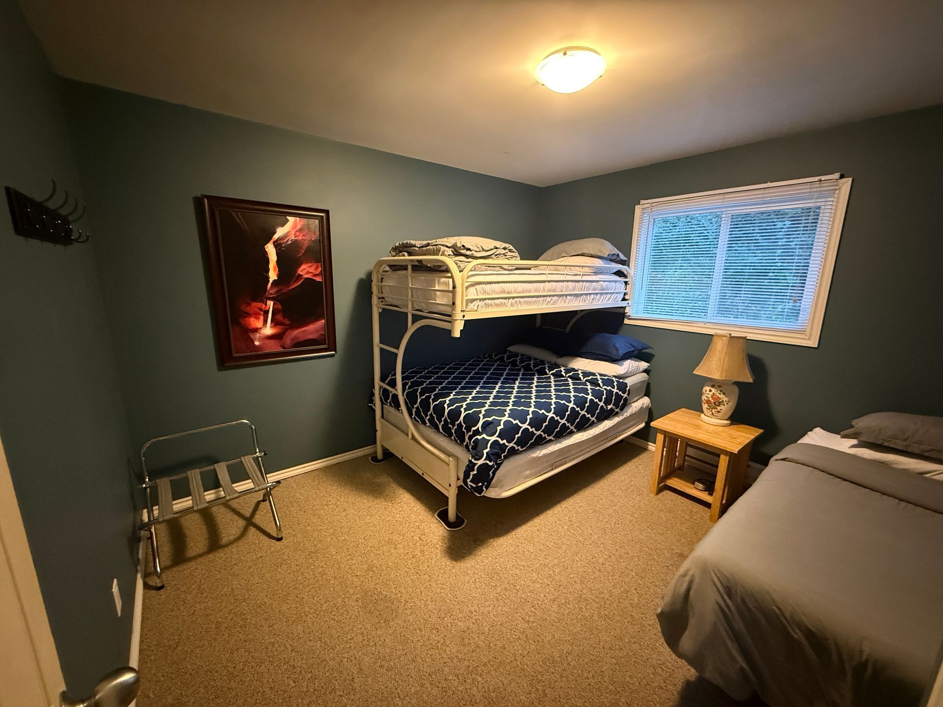 A bedroom with dark blue walls, a white bunk bed, a twin bed, and a small wooden nightstand with a lamp.