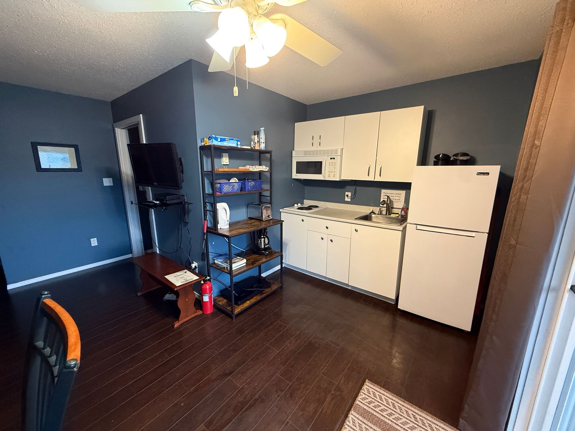 A kitchen area with white cabinets, a refrigerator, and a dark shelving unit against dark blue walls on wood floors.