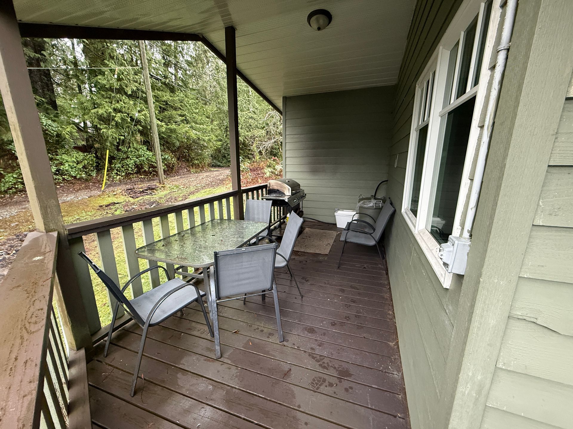 A patio deck with a glass-topped table and chairs, overlooking a wooded area on an overcast day.
