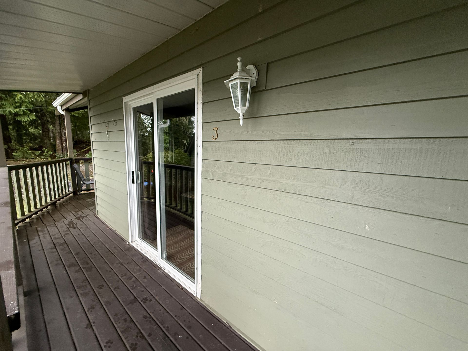 A green-sided house exterior featuring a wooden porch, a sliding glass door, and a white outdoor wall lamp.