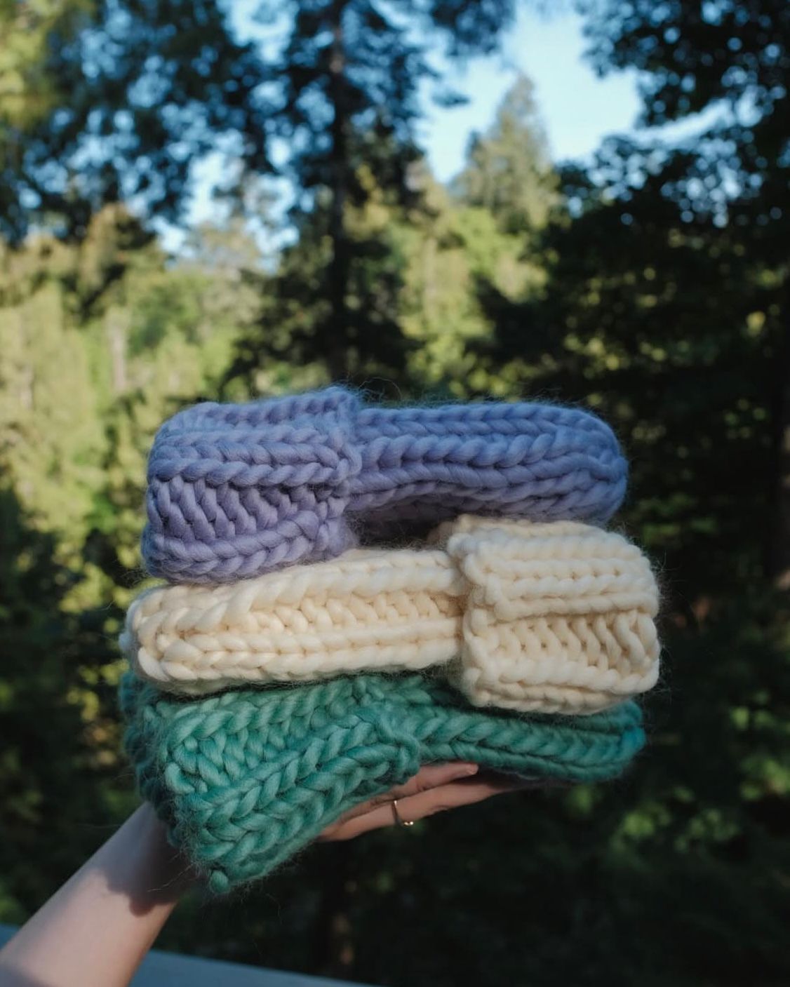 A hand holds a stack of three chunky, cable-knit beanies in lavender, cream, and teal against a background of trees.