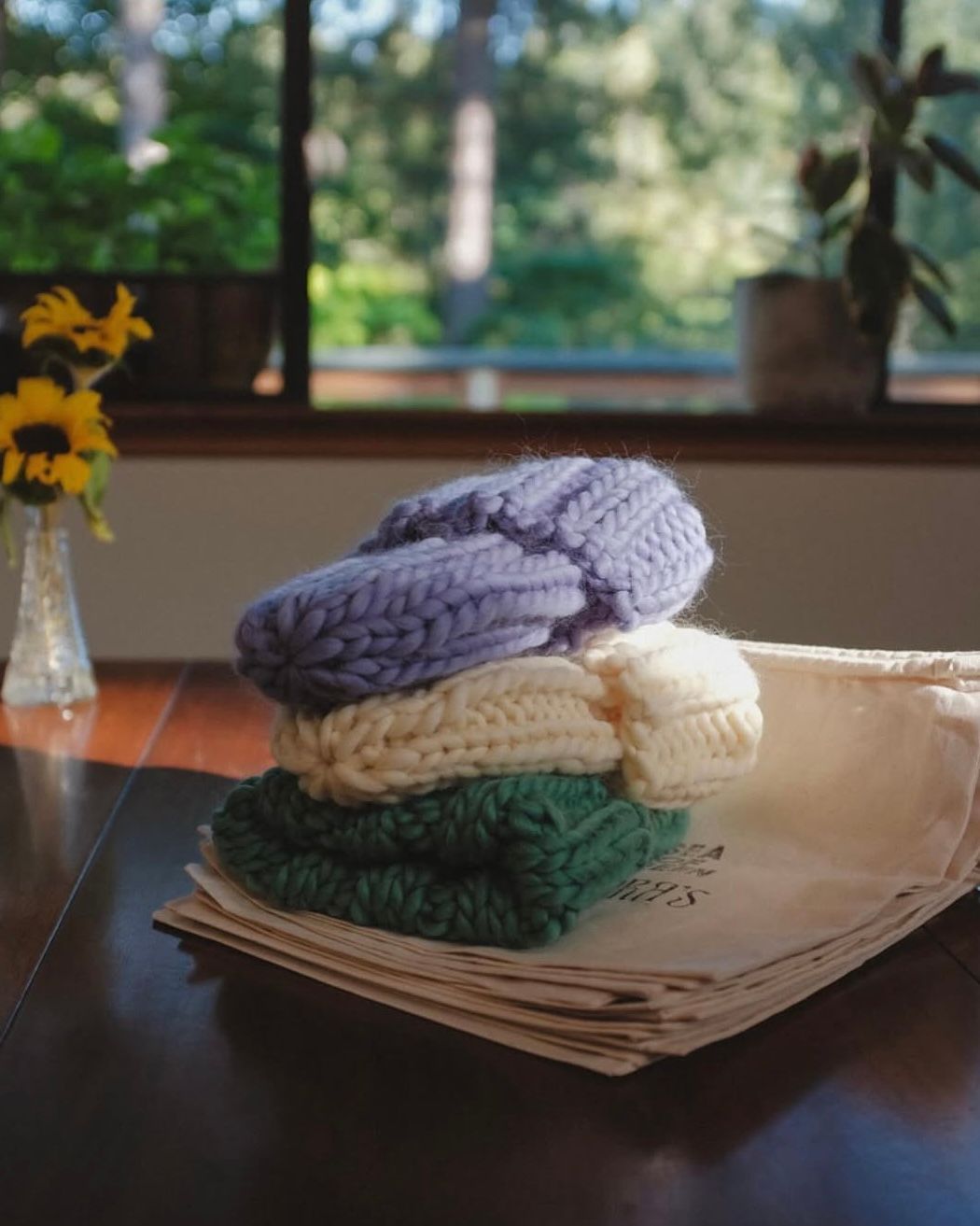 A stack of three chunky knit hats in blue, cream, and green resting on paper near a vase of sunflowers by a window.