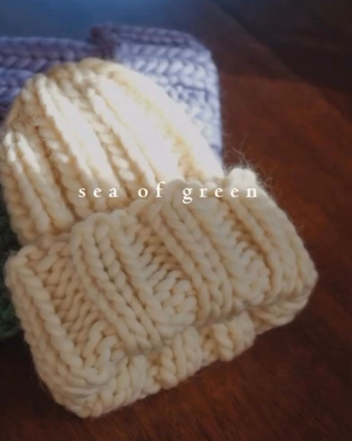 A cream-colored, chunky knit beanie with a folded brim sits on a wooden surface, with a blurred purple hat in the back.