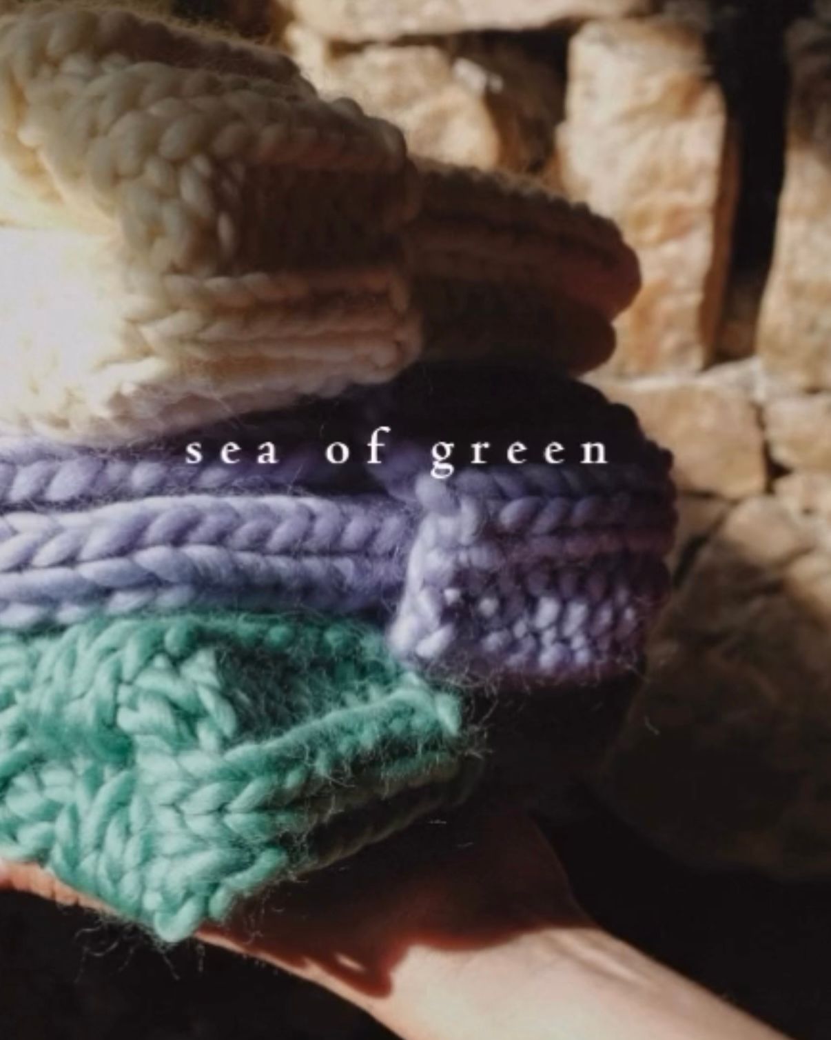 A hand holds a stack of three chunky knit beanies in cream, lavender, and mint green, against a rustic stone background.