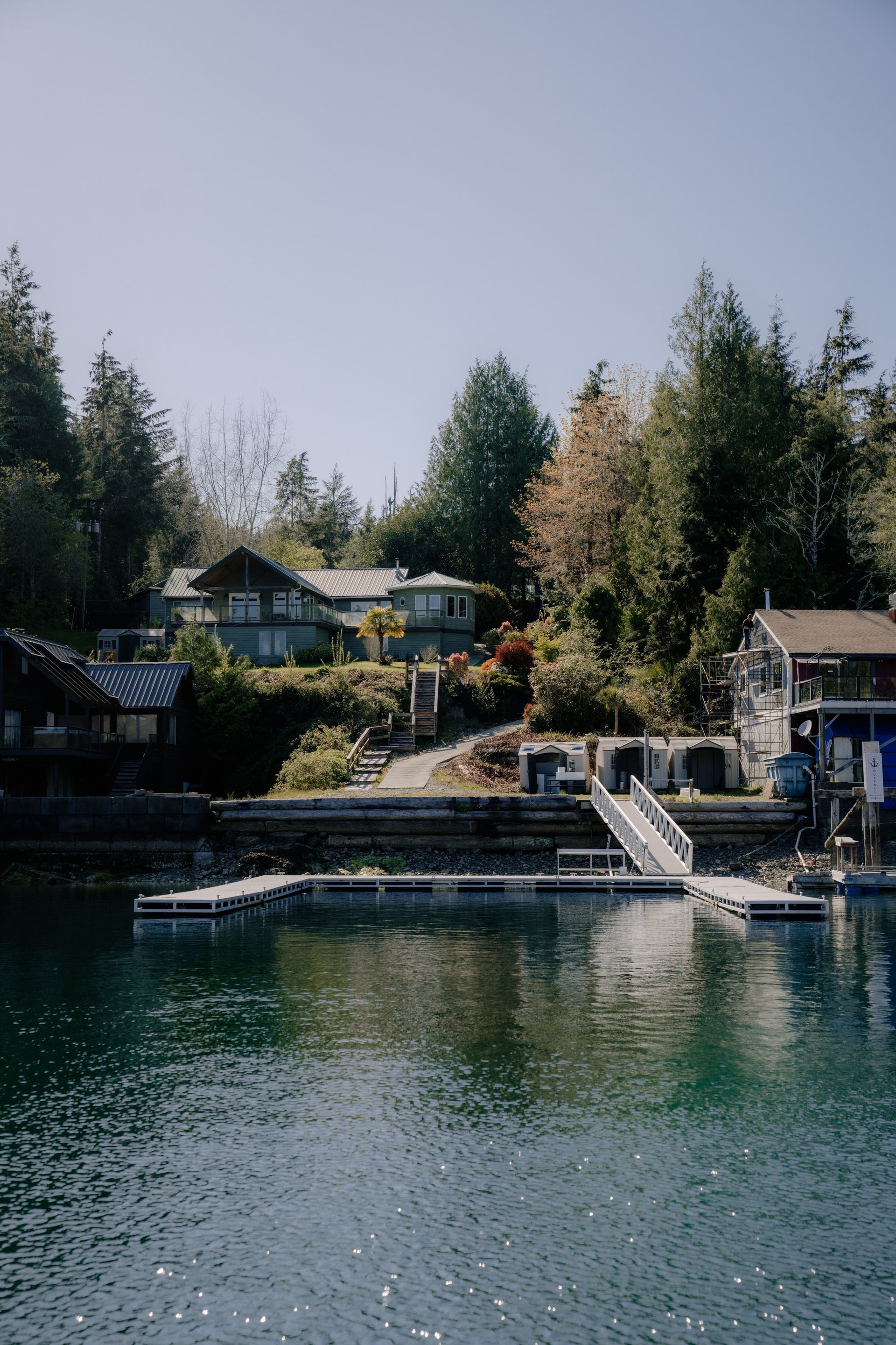 A house perched on a wooded hillside overlooking a calm, deep-blue bay with a wooden dock and ramp in the foreground.