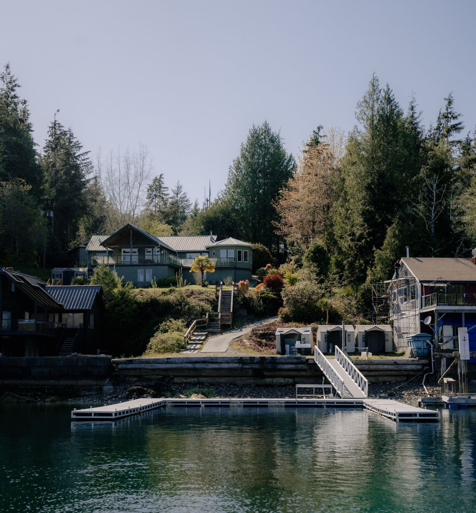 A house on a hillside overlooks a calm waterfront, featuring a wooden dock and a ramp extending into the water.
