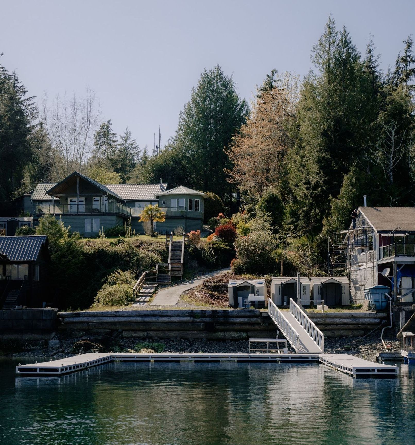 A multi-level house nestled in a wooded hillside above a floating U-shaped dock on a calm, deep blue waterway.