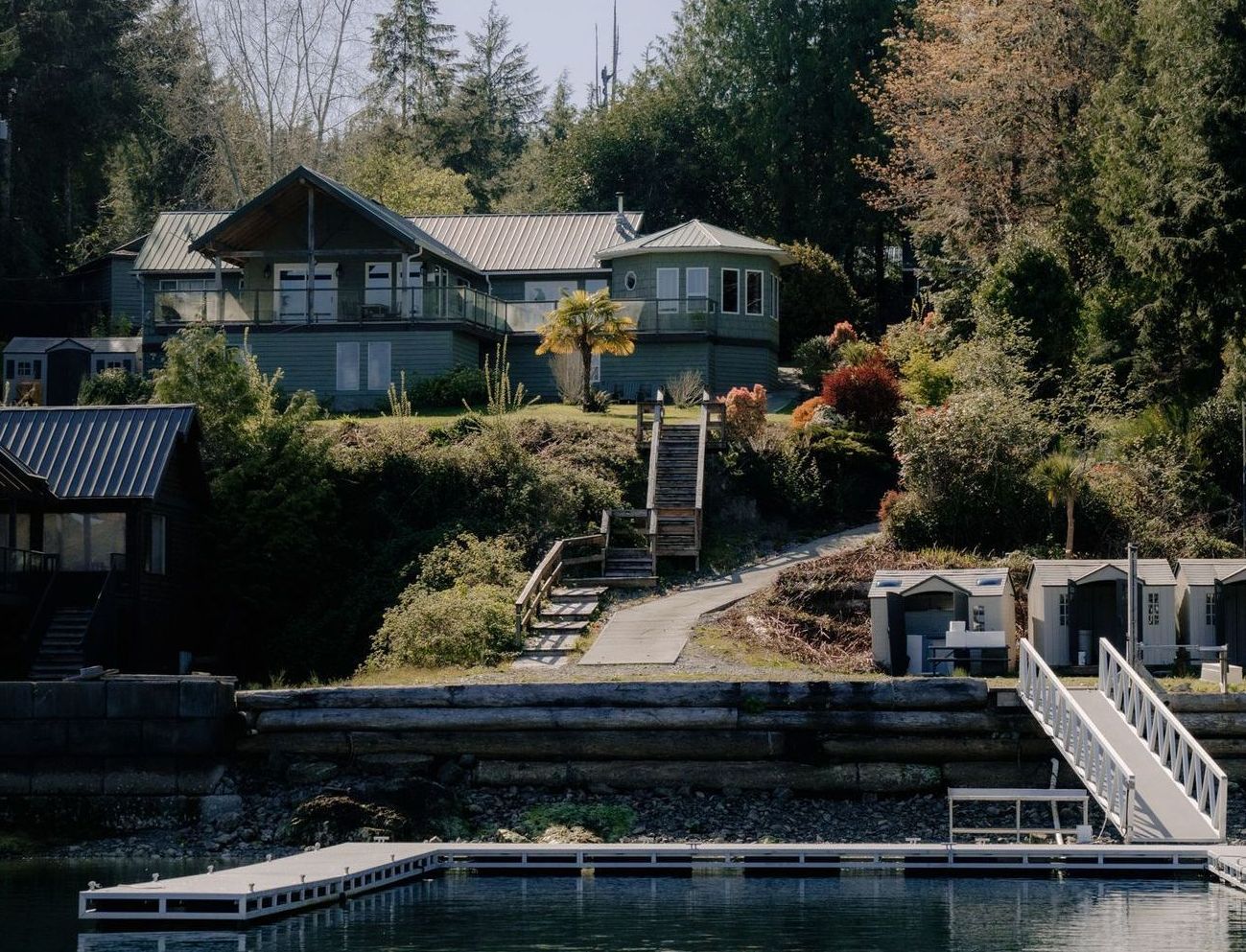 A house on a hillside with stairs leading down to a wooden retaining wall and a floating dock on the water.