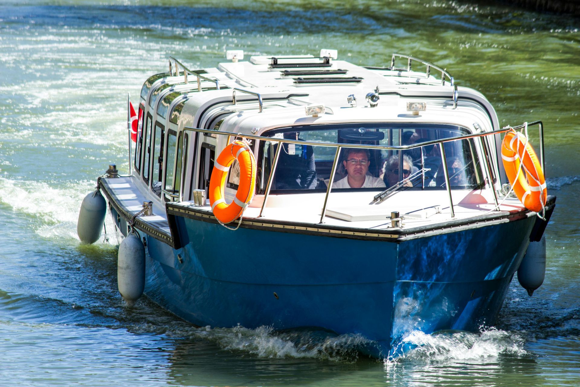A blue and white tour boat cruises on calm water, featuring orange life preservers attached to its front railings.