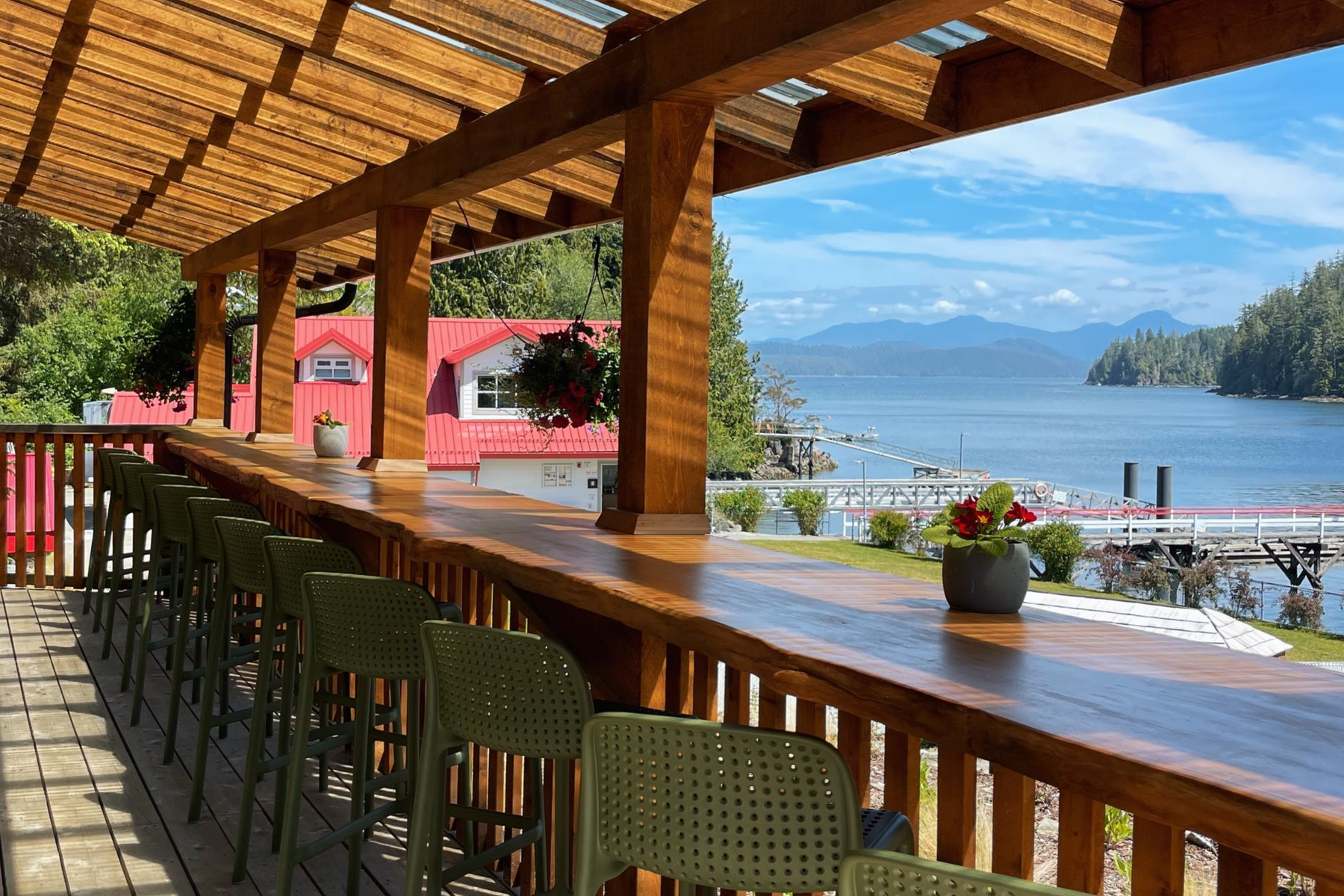 An outdoor wooden bar counter overlooking a calm lake, lush forest, and distant mountains on a sunny day.