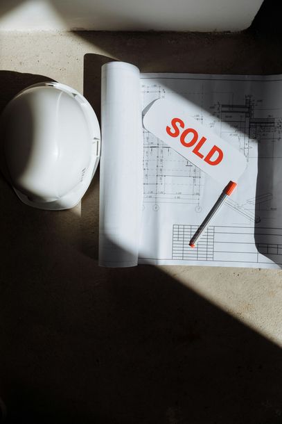 A hard hat , blueprints , and a sold sign on a table.