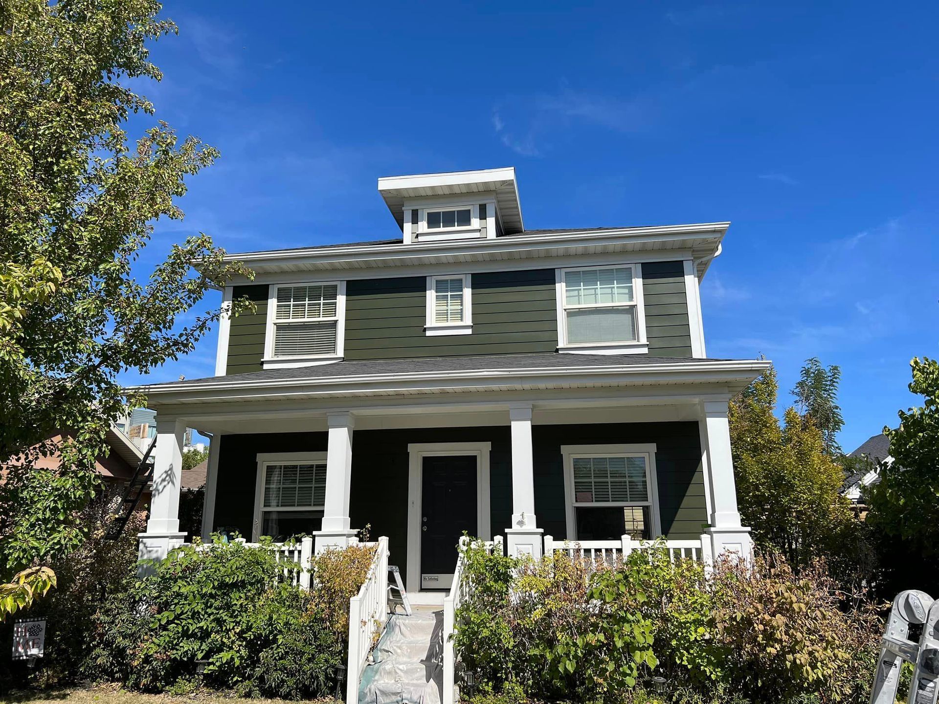 A large house with a green siding and white trim is surrounded by trees and bushes.