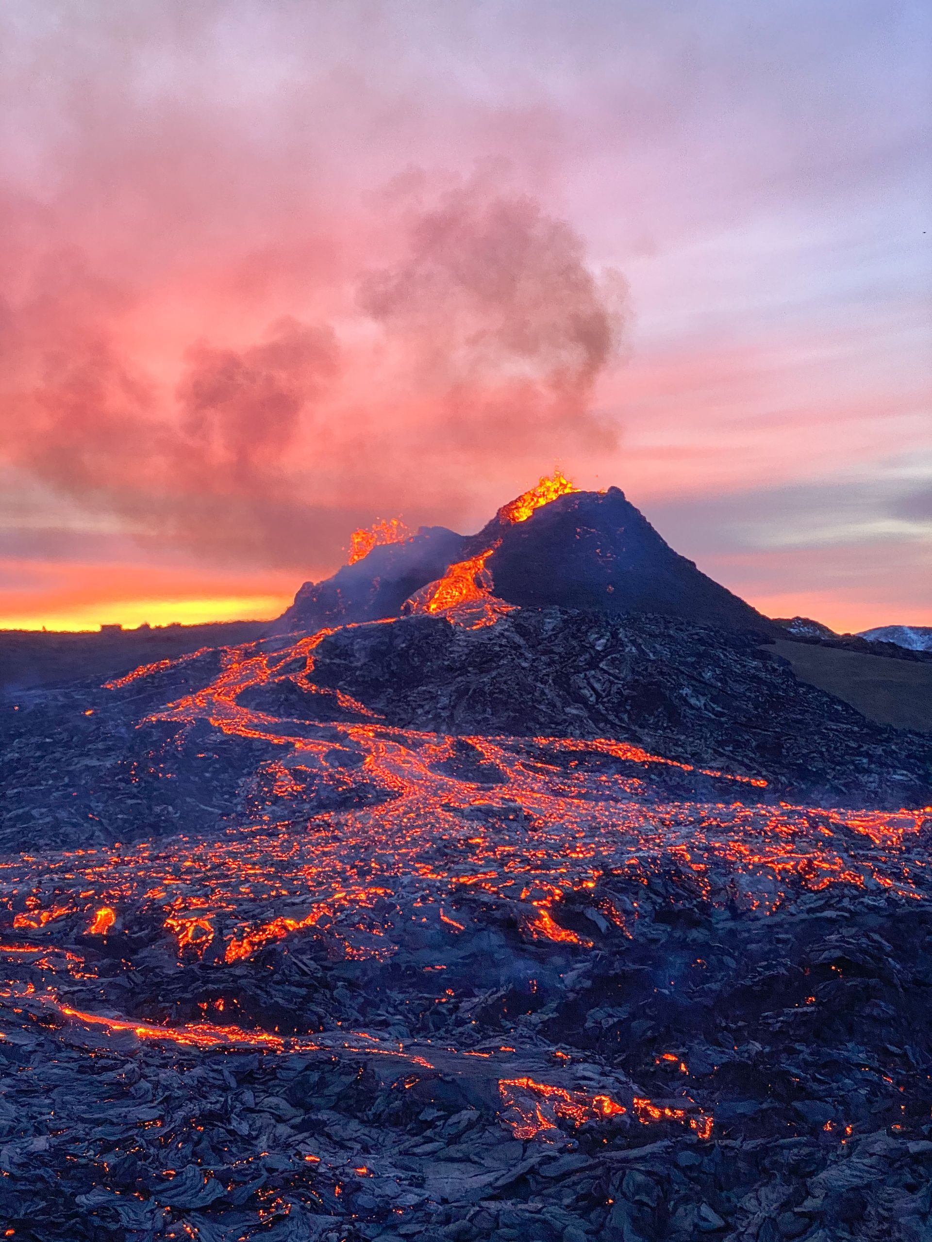 A volcanic crater with calm lava pouring down its side and a pink sunrise in the background.