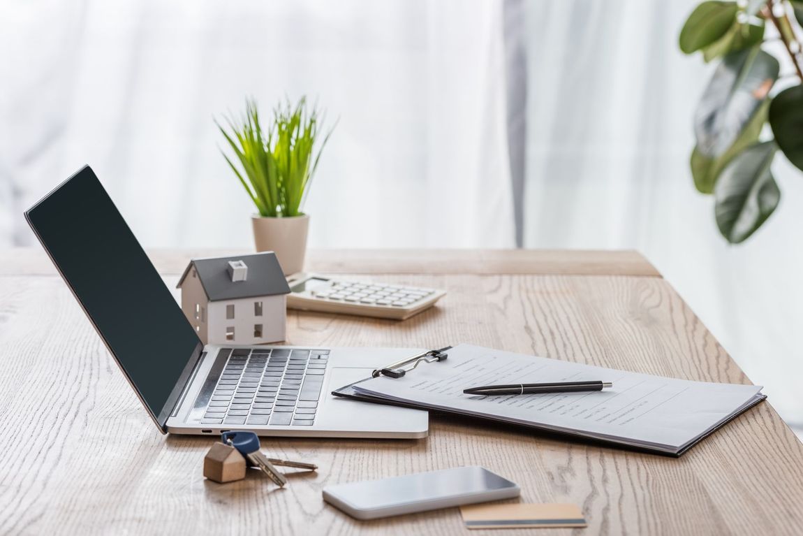 A wooden desk with a laptop , keys , a cell phone , and a model house.