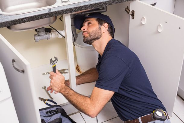 Plumber working under a kitchen sink, using a wrench. Wearing blue shirt and hat. Plumber working under a kitchen sink, using a wrench. Wearing blue shirt and hat.