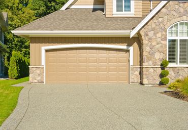A beige garage door with a curved top trim, set in a house with stone siding and a paved driveway under a sunny sky.