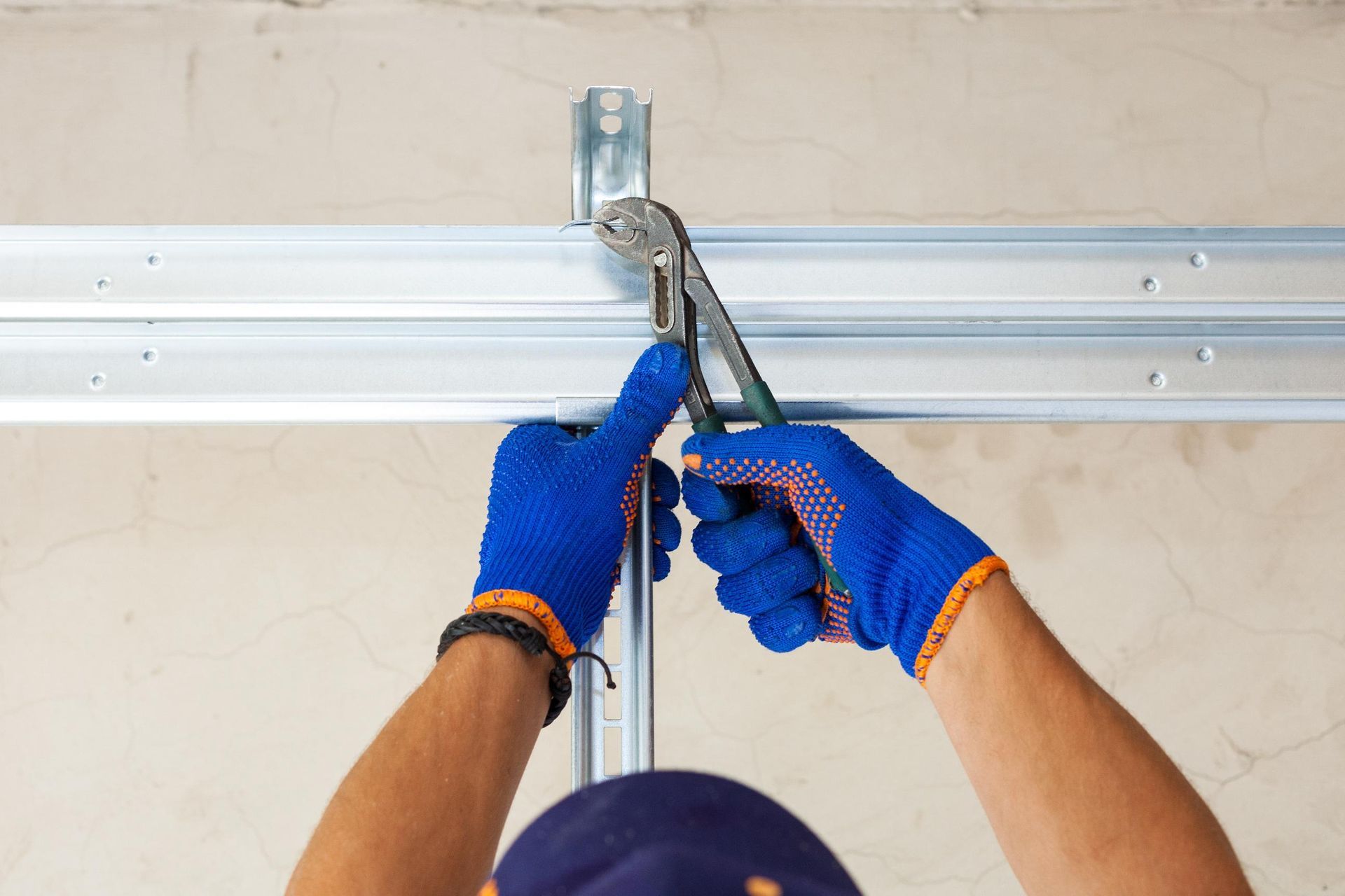 Hands in blue work gloves use pliers to attach a metal profile to a ceiling structure.