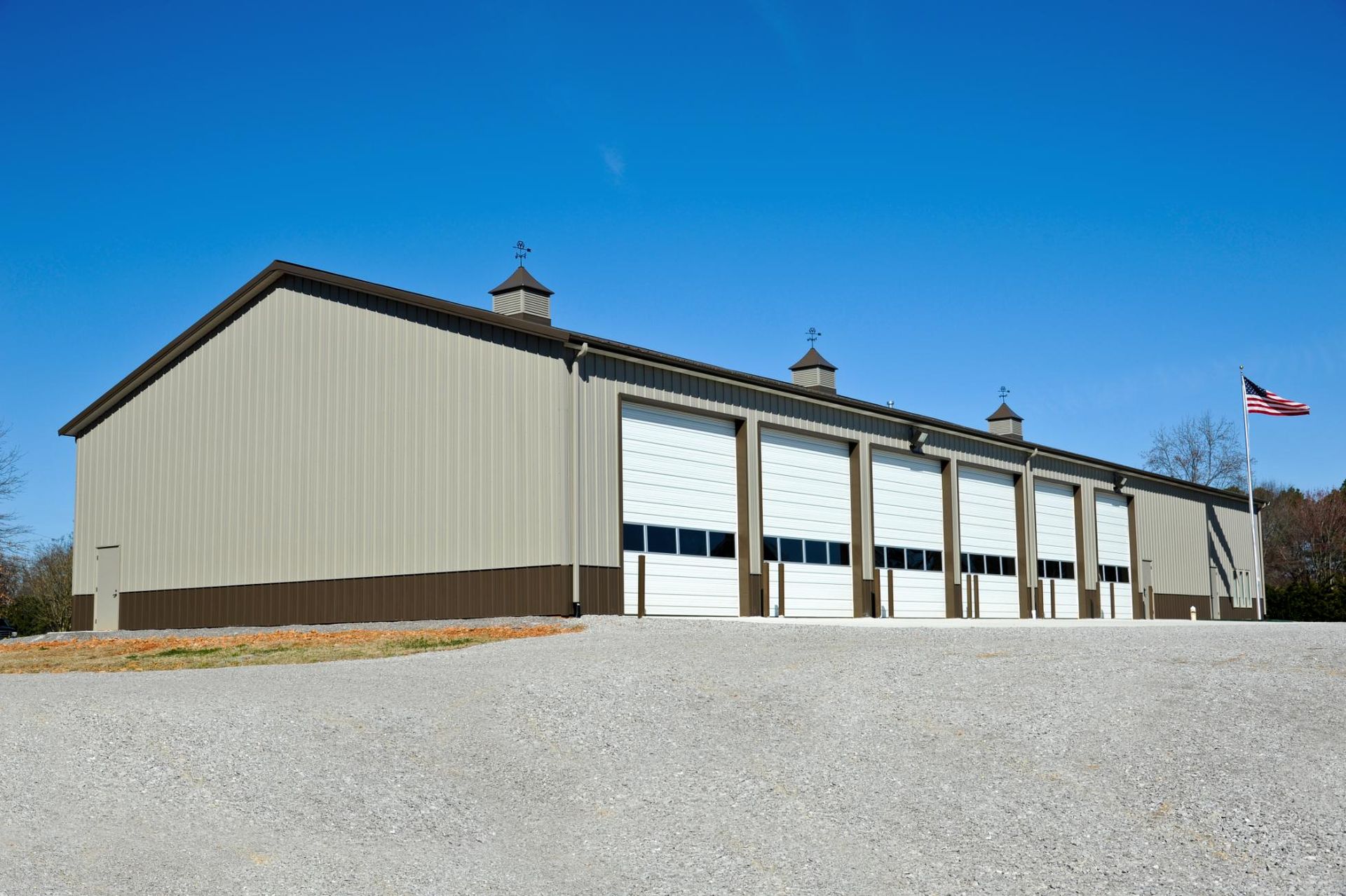 A tan metal building with five white garage doors and a US flag in a gravel parking lot under a clear blue sky.