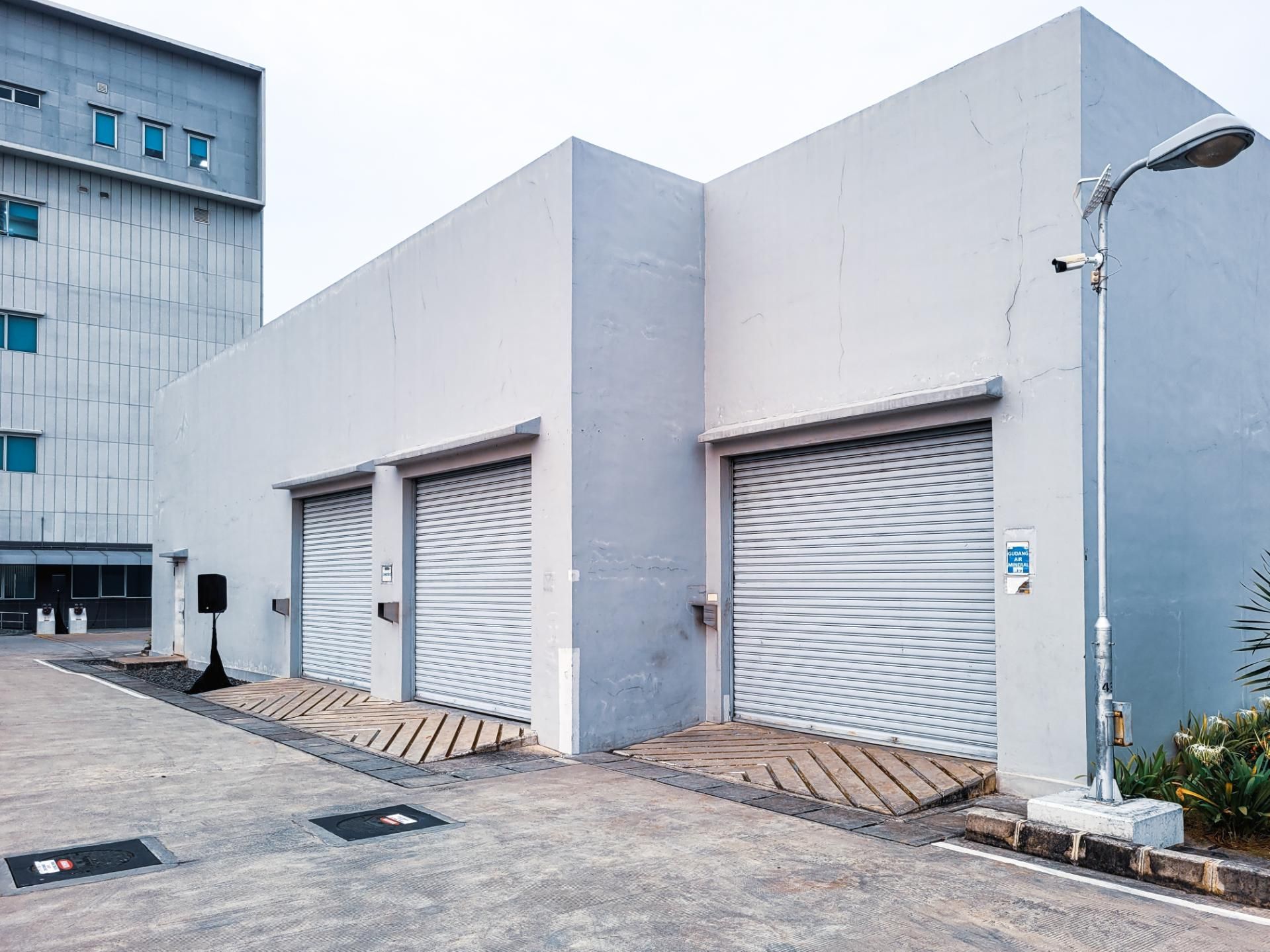A modern grey building exterior with three closed metal roll-up garage doors, a light pole, and a paved lot.