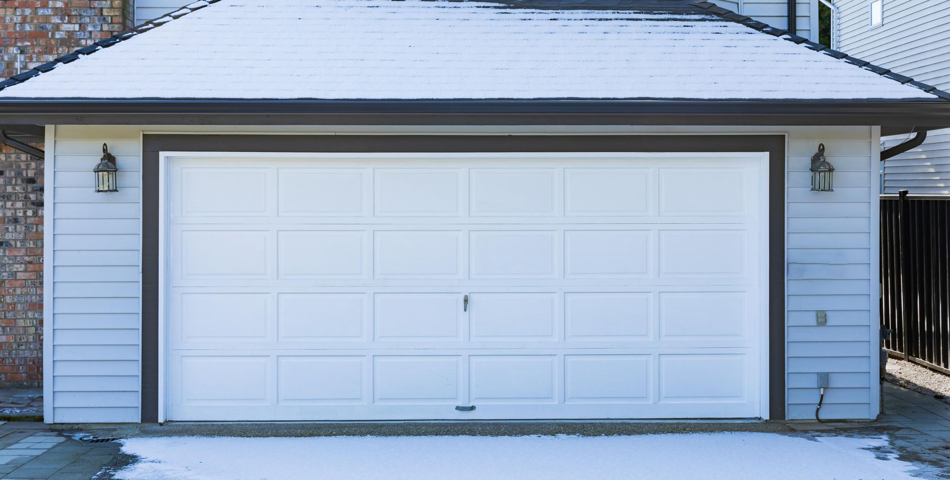 A white garage door with rectangular panels is set in a light grey house exterior with snowy ground under the roof eaves.