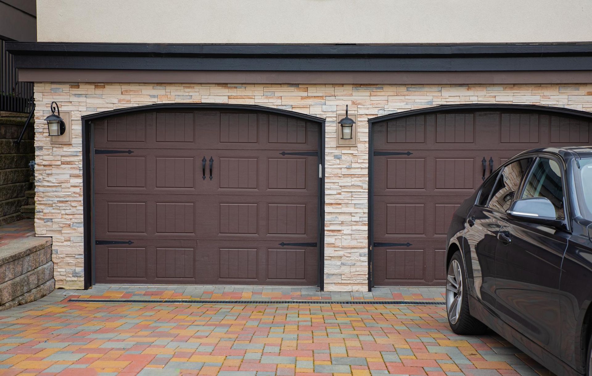 Two dark brown carriage-style garage doors with iron hardware set in a stone wall above a colorful paver driveway.