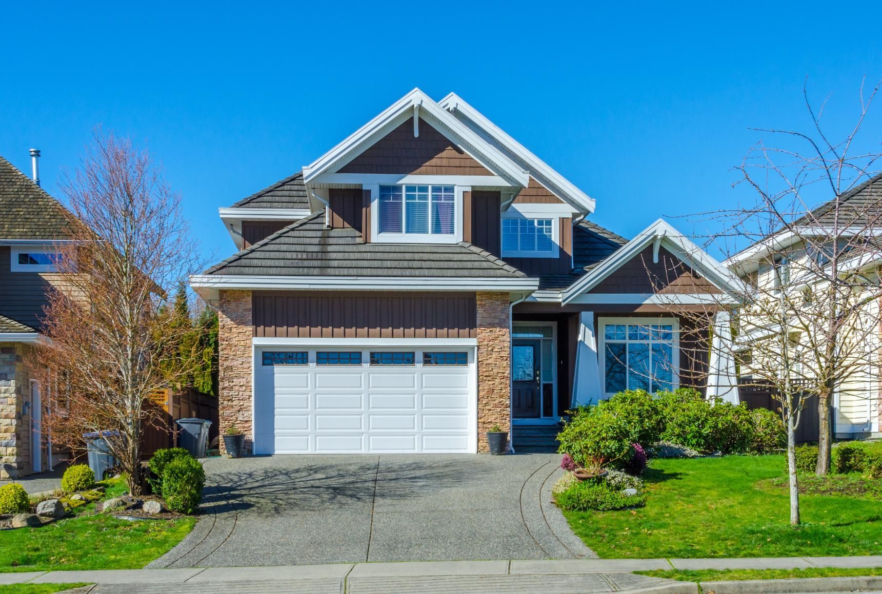 A two-story suburban house with brown siding, stone accents, a white garage door, and a paved driveway under a blue sky.