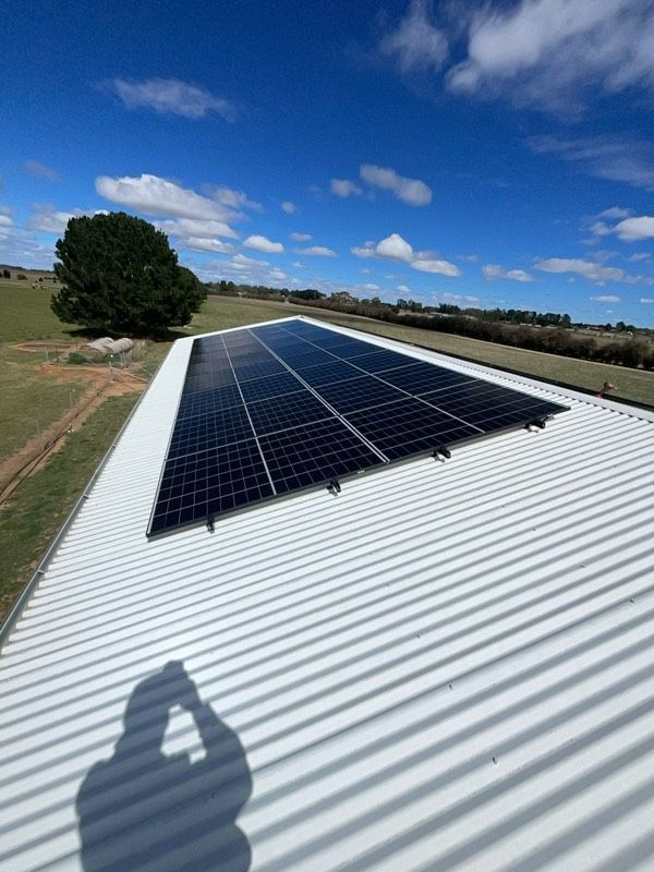 Solar panels on a white corrugated roof under a blue sky with a shadow.