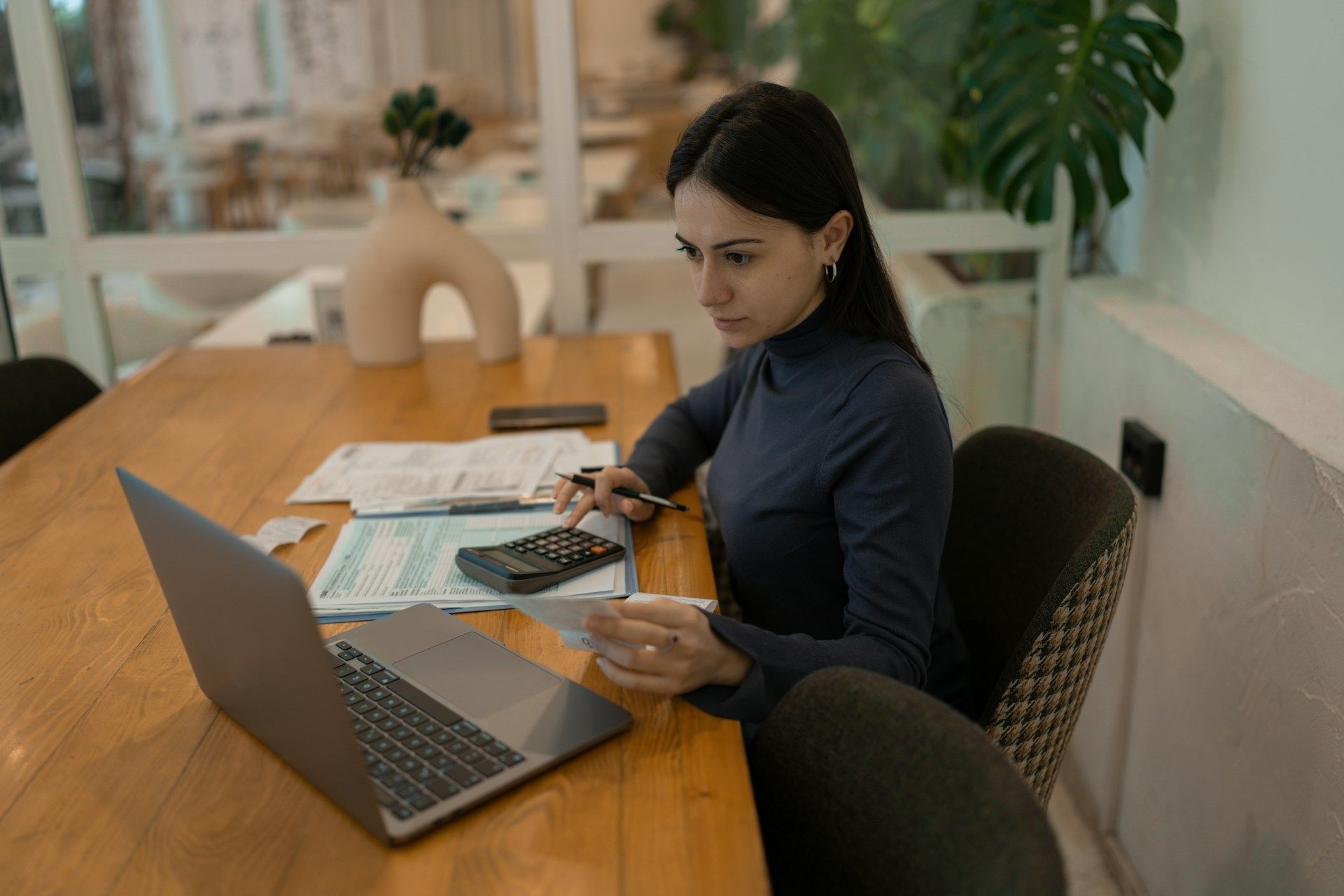 Woman using laptop, calculator, and paperwork at wooden table in a well-lit room.