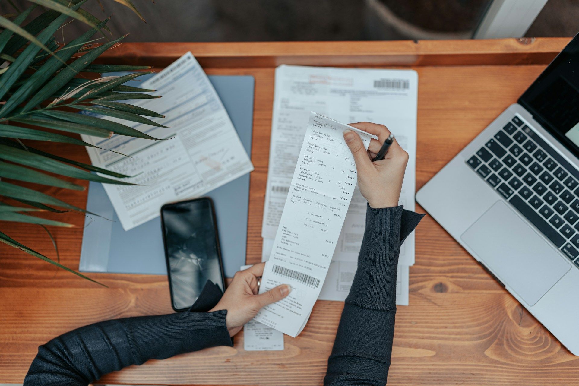 Person reviews long receipt, writing with pen, with laptop, phone, and papers on wooden desk.