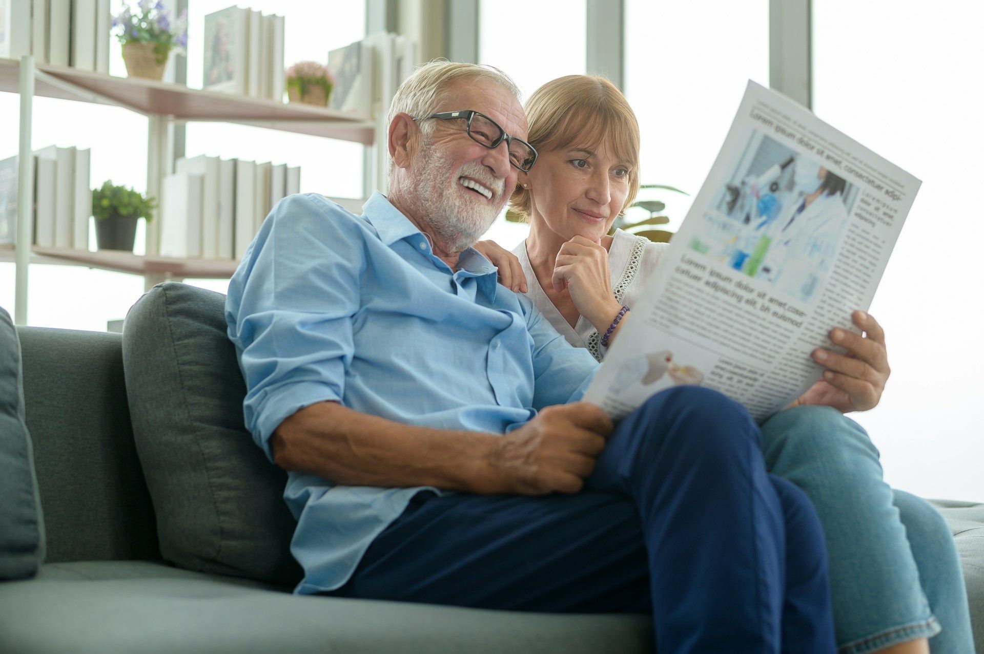 Smiling older couple reading newspaper together on a gray sofa in a bright room.