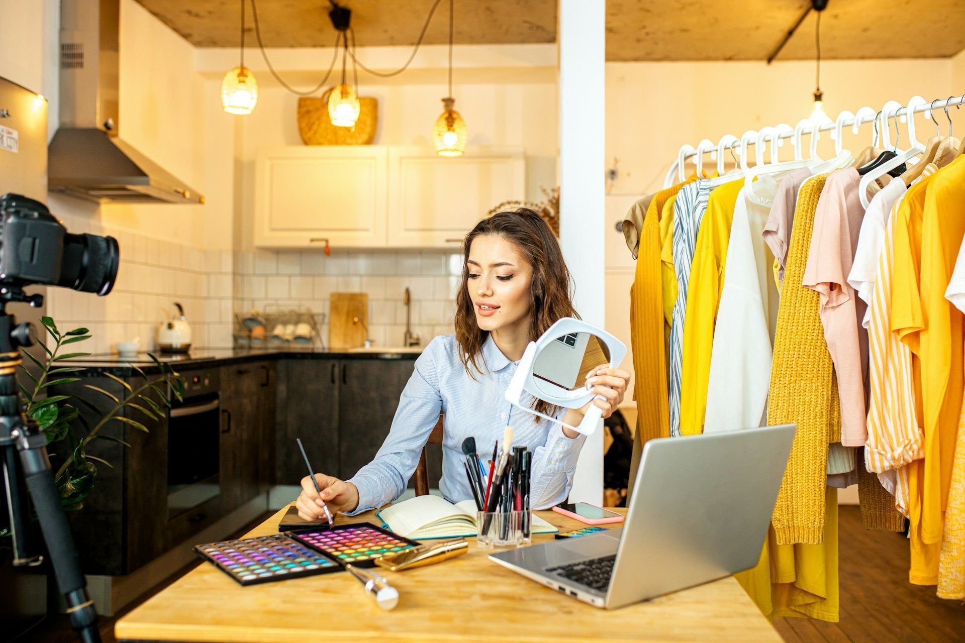 Woman filming makeup tutorial in a kitchen studio with laptop and clothing rack, holding ring light and palette.