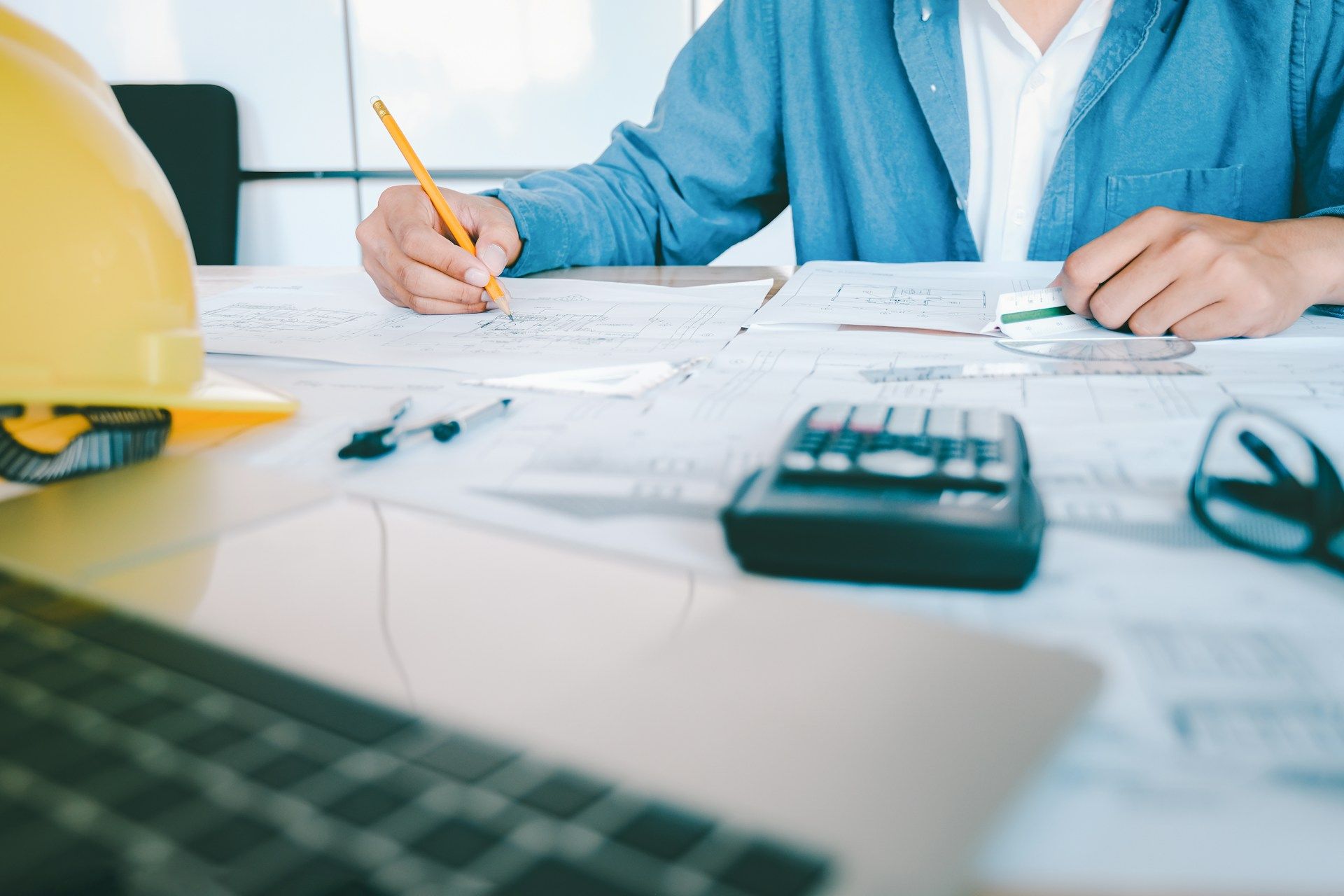 Architect working on blueprints at a desk, yellow hard hat, laptop, calculator, and glasses visible.