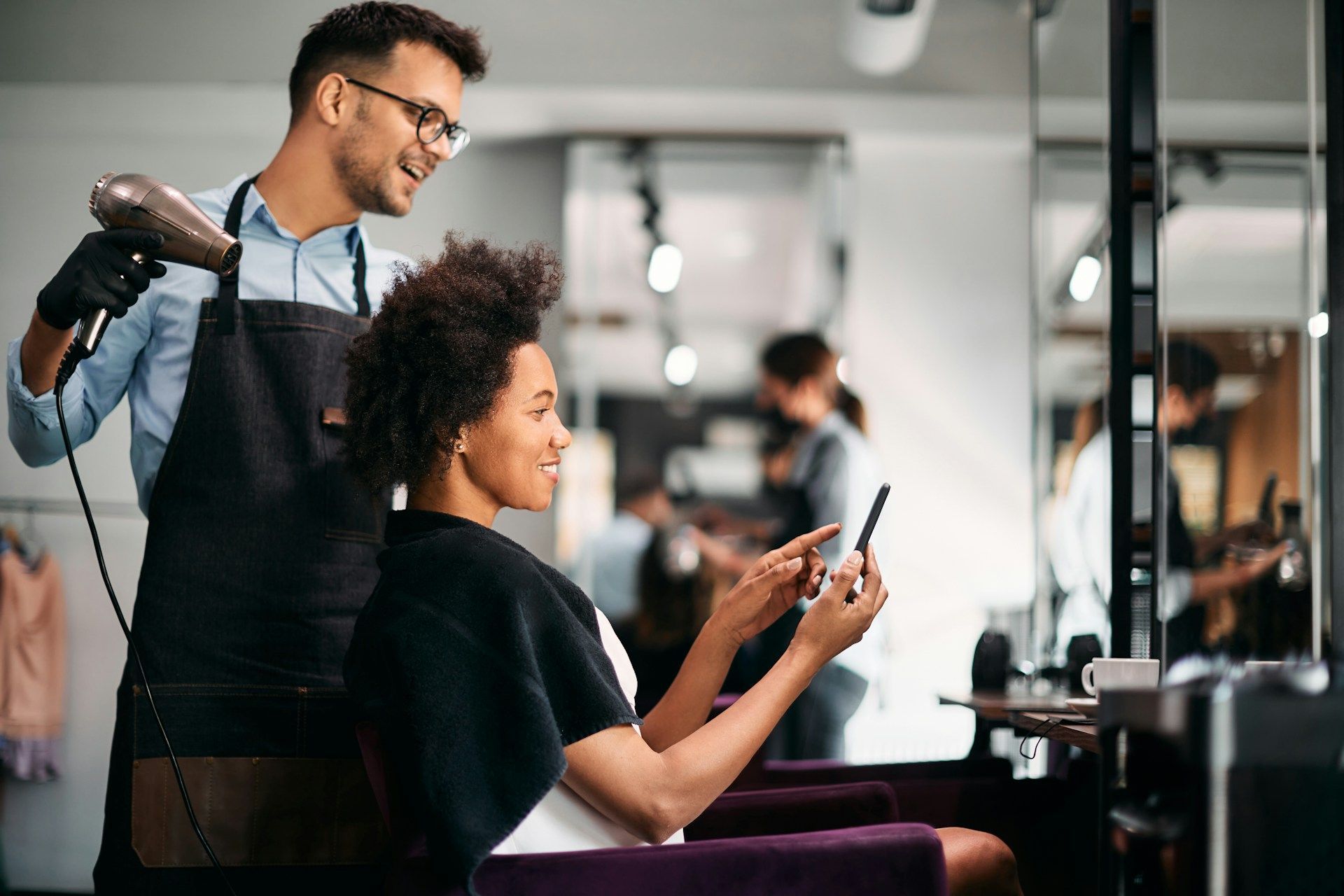 Hairdresser drying client's hair, client looks at phone in a salon.