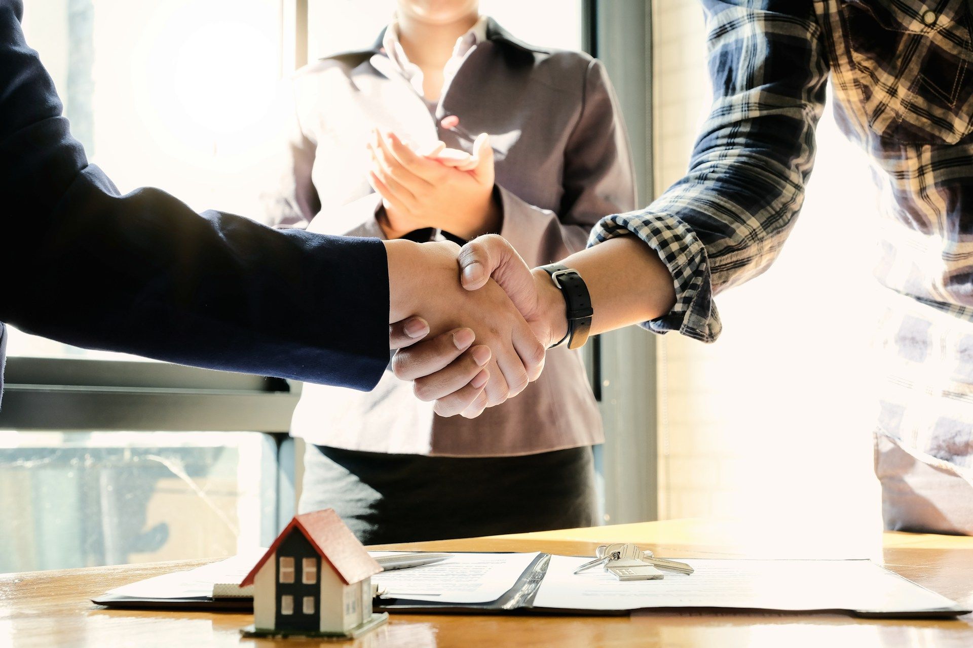 Two people shaking hands over a table with a small house model and documents; a third person claps in the background.
