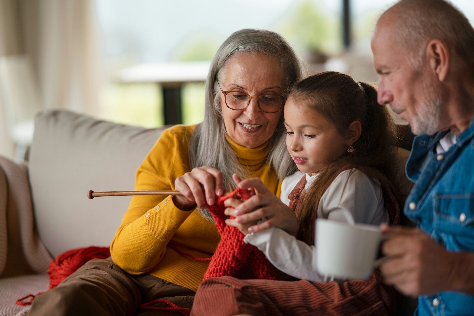 Grandmother teaches granddaughter to knit on a couch, grandfather watches, holding a mug.