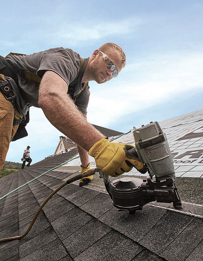 Roofer using a nail gun on a shingled roof, wearing safety glasses and gloves, working outdoors.