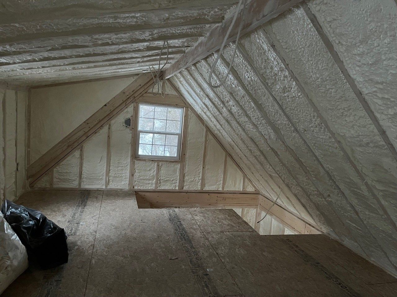 Attic space with spray foam insulation on walls and rafters, unfinished wooden floor, small window.