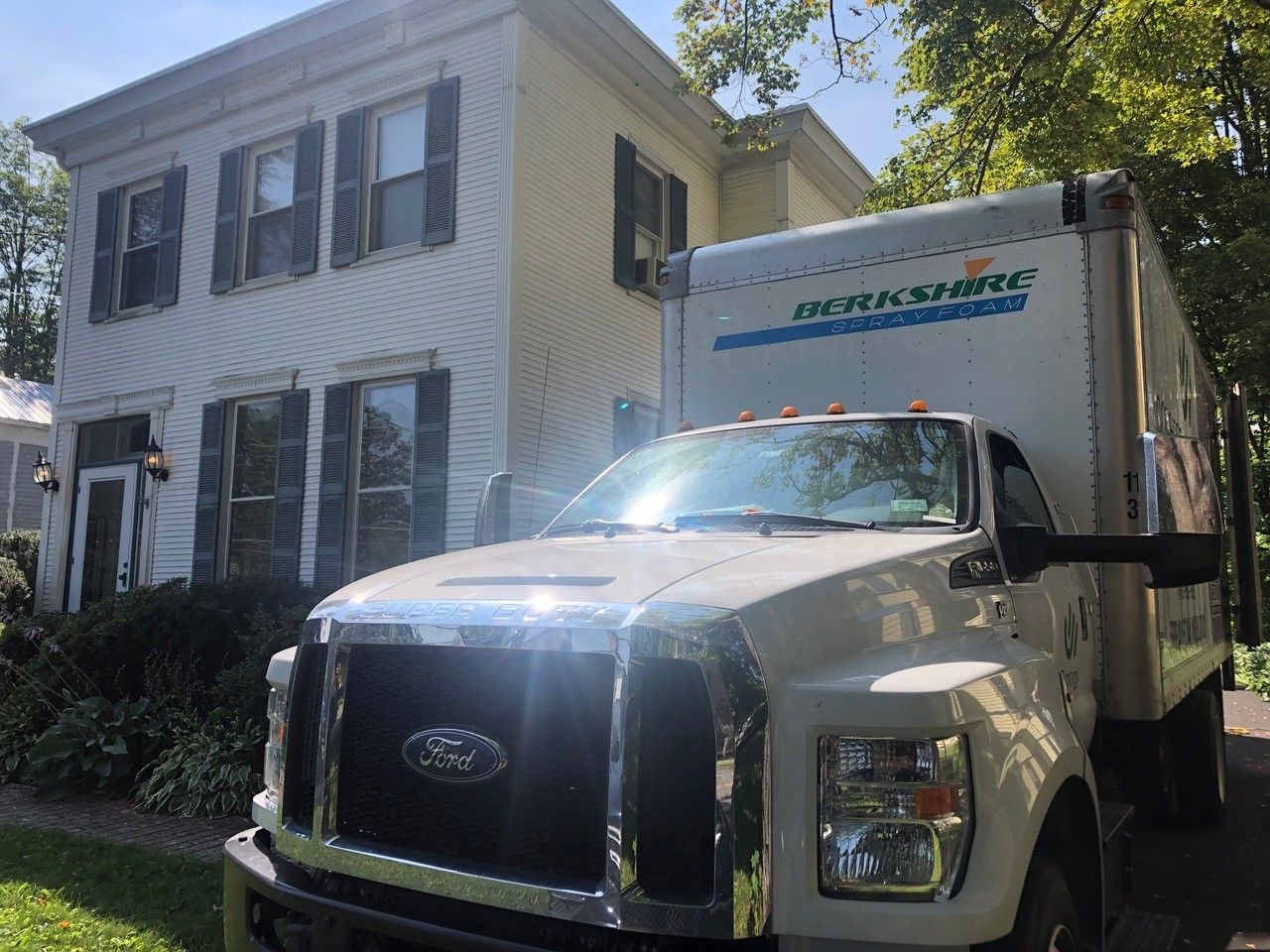 White moving truck parked in front of a two-story white house with black shutters on a sunny day.