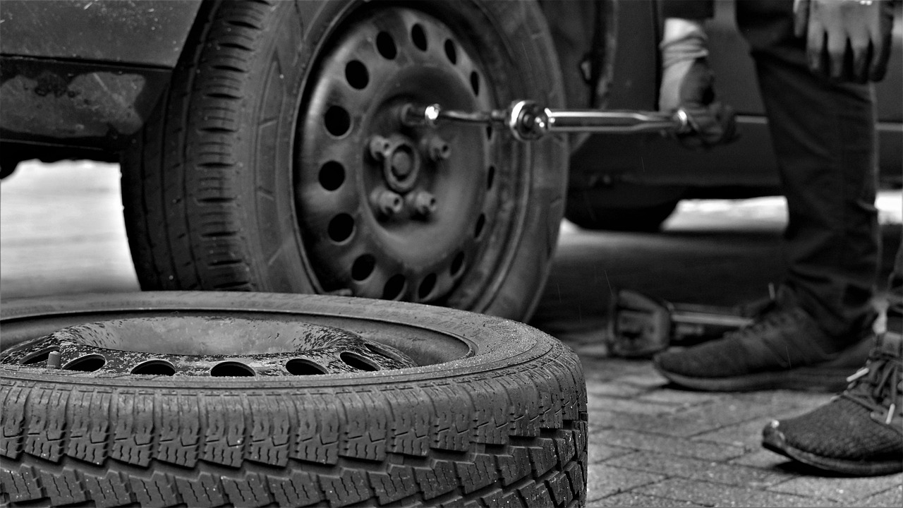 Person changing a car tire beside a spare tire on a sidewalk, in black and white