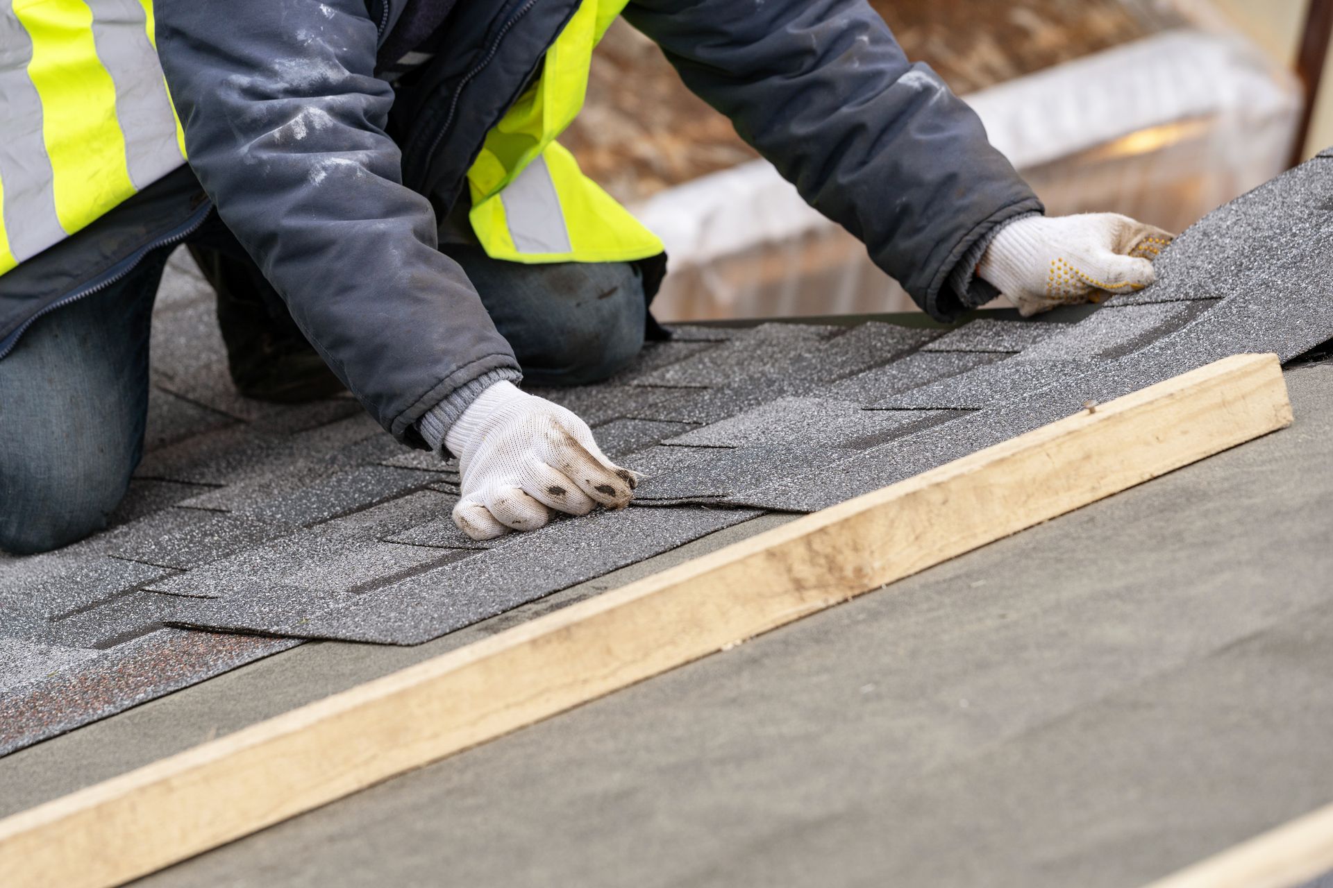 Person in safety vest installing gray asphalt roof shingles.
