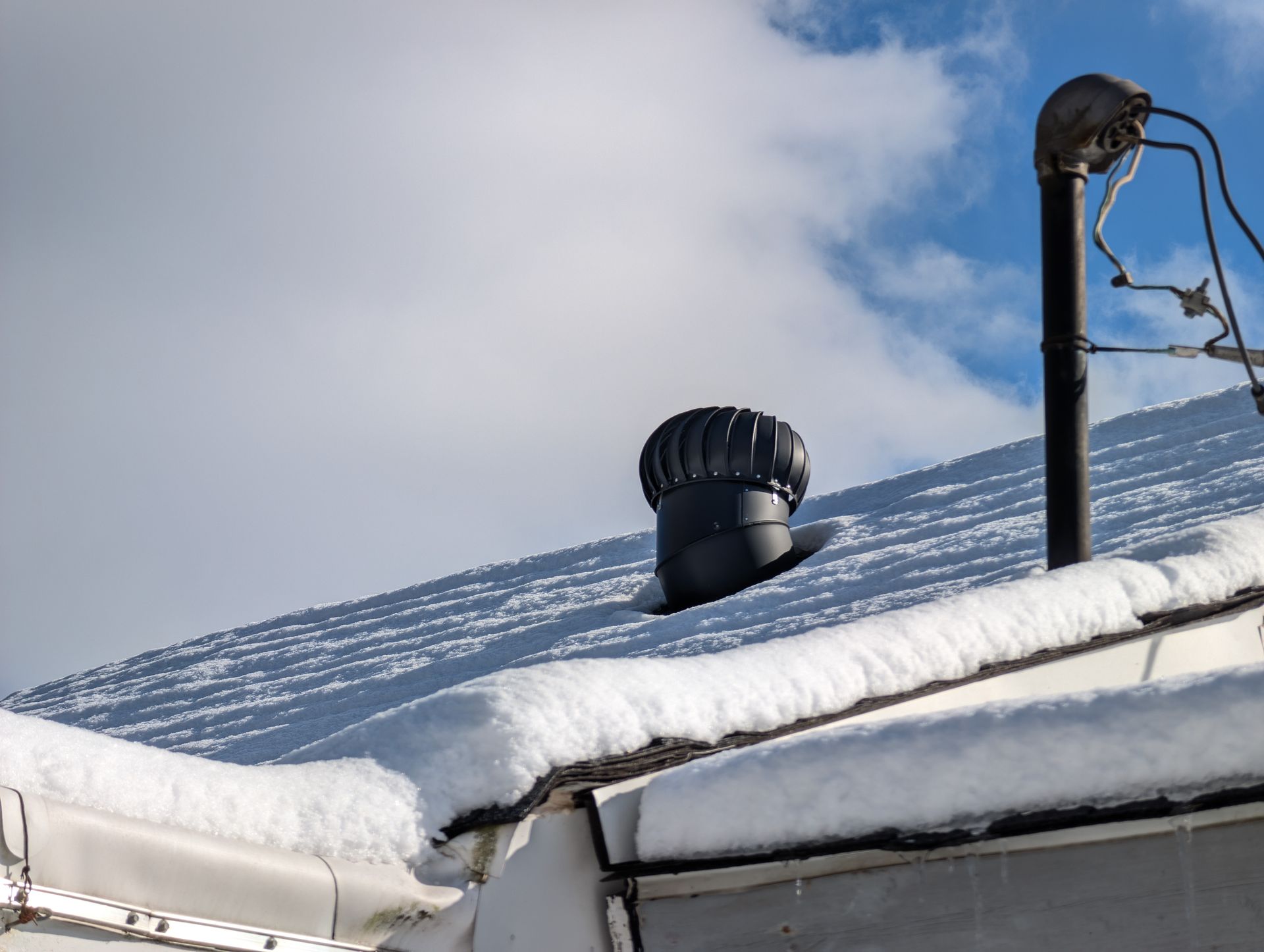 Snow-covered roof with a black vent and chimney against a cloudy blue sky.