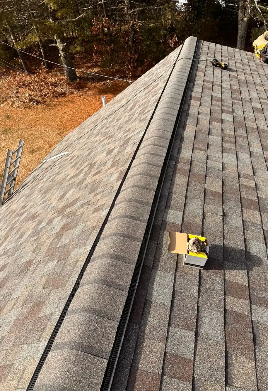 View of a brown asphalt shingle roof with the ridge cap being installed. A cardboard box and tools are placed on the roof.