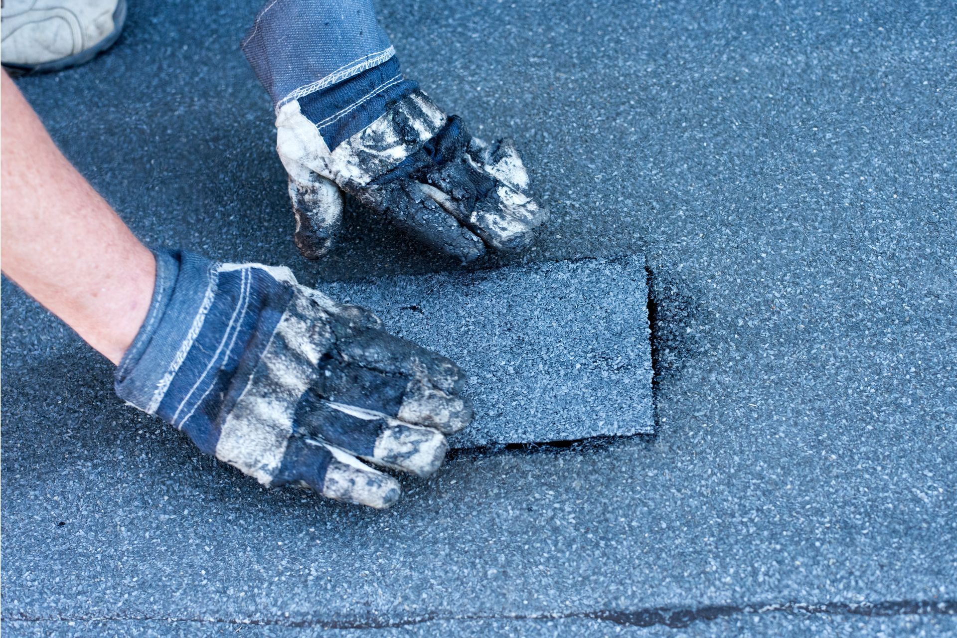 Gloved hands holding a torn piece of blue-gray roofing material on a roof surface