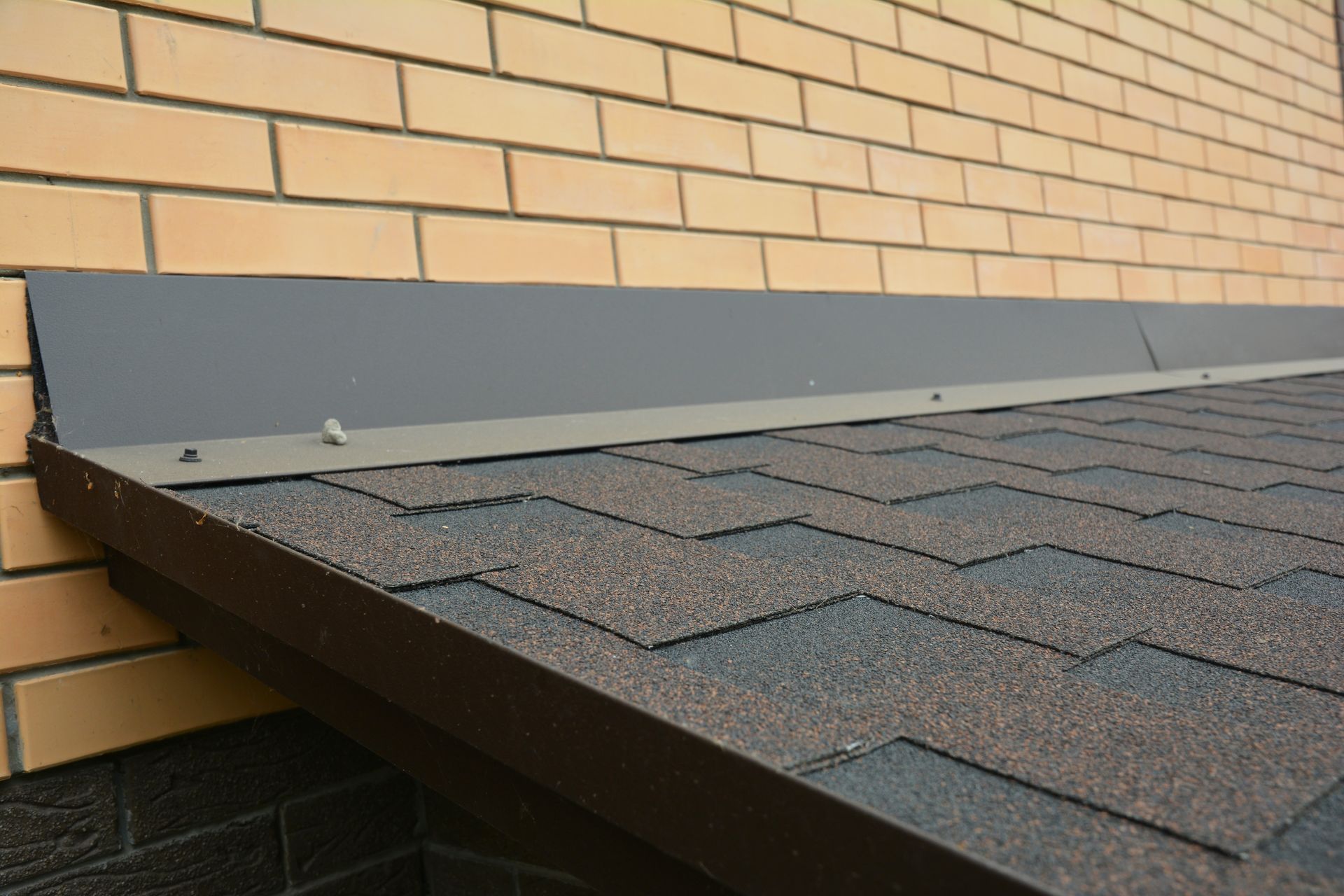 Close-up of a shingled roof edge against a brick wall with flashing and gray metal trim.