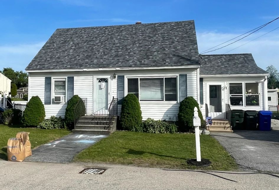 A white house with a grey roof and blue shutters, with green lawn and small bushes in front.